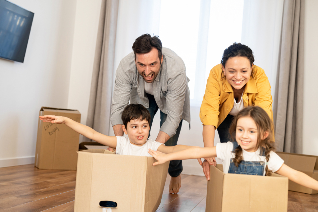 Multiracial family of four have fun in new house, parents riding kids in cardboard boxes in empty living room, happy children spreading arms and laughing